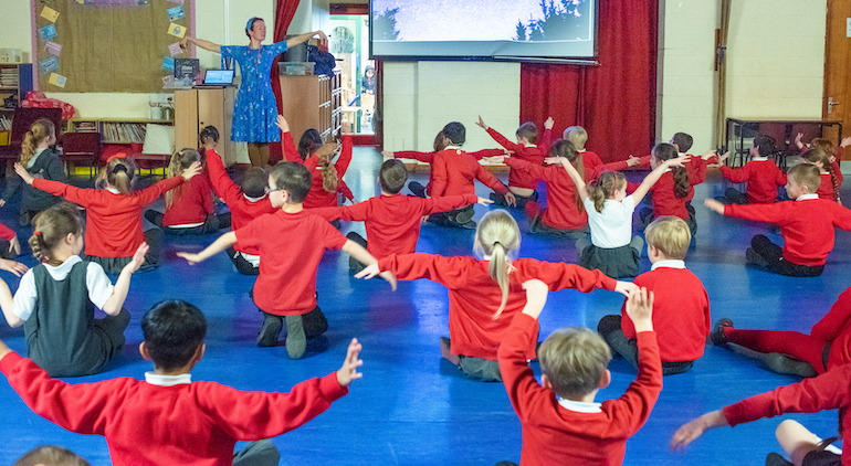 pupils seated on the floor in red uniform are guided through yoga session by a grown up at the front of the hall