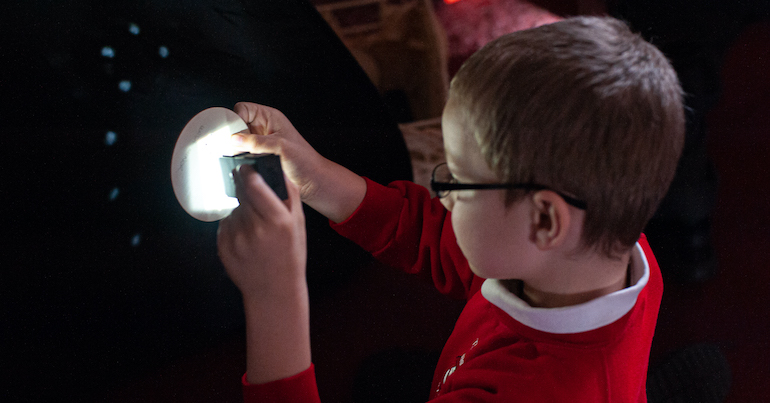A boy uses a torch in a dark room to explore a map of the constellations