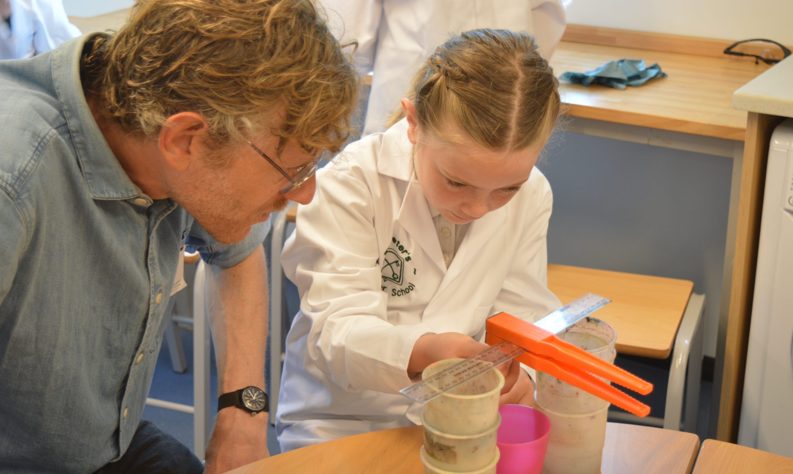 a female pupil in a white lab coat is conducting a science investigation whilst a grown up looks on closely