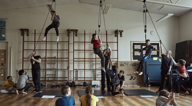 children in a school hall are lifted into the air with harnesses and pulley systems as they explore forces