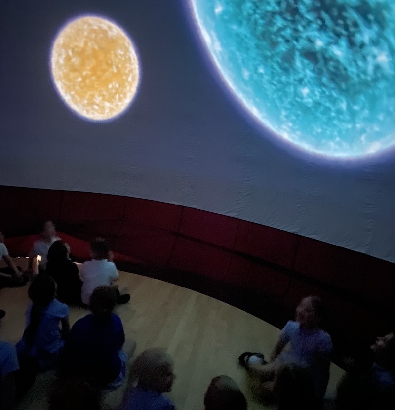 two large images of planets in a dark space with children seated on the floor looking at them