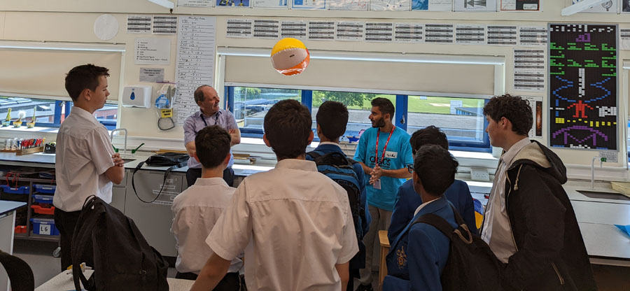 A group of students watch a science experiment that shows a beach ball floating in the air. A teacher and demonstrataor are in front of the group.