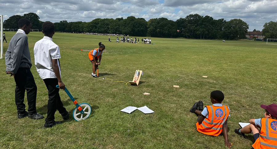 students on a field watching one student launch a rocket - two students stood with a measuring wheel; two students sat on floor in orange hi vis jackets